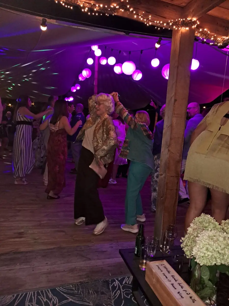 Gasten van alle leeftijden dansen op een houten dansvloer onder paarse lampionnen en lichtsnoeren, een oudere dame in een gouden jasje danst vooraan