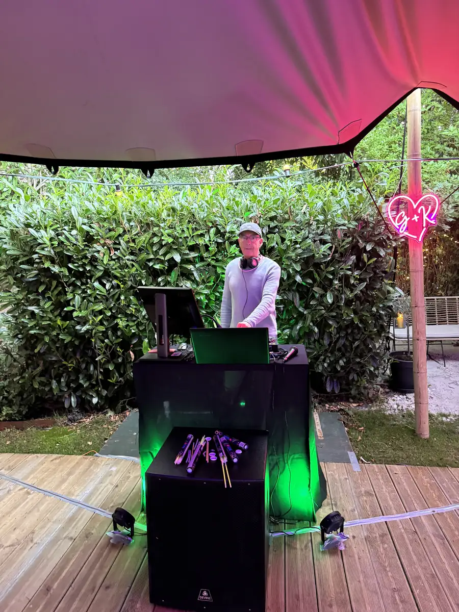 DJ Paul van Sun Groove Music achter zijn DJ-booth onder een rood verlicht zeildoek in een tuin, met een roze neonhart en groene sfeerverlichting, vlak voor een feest