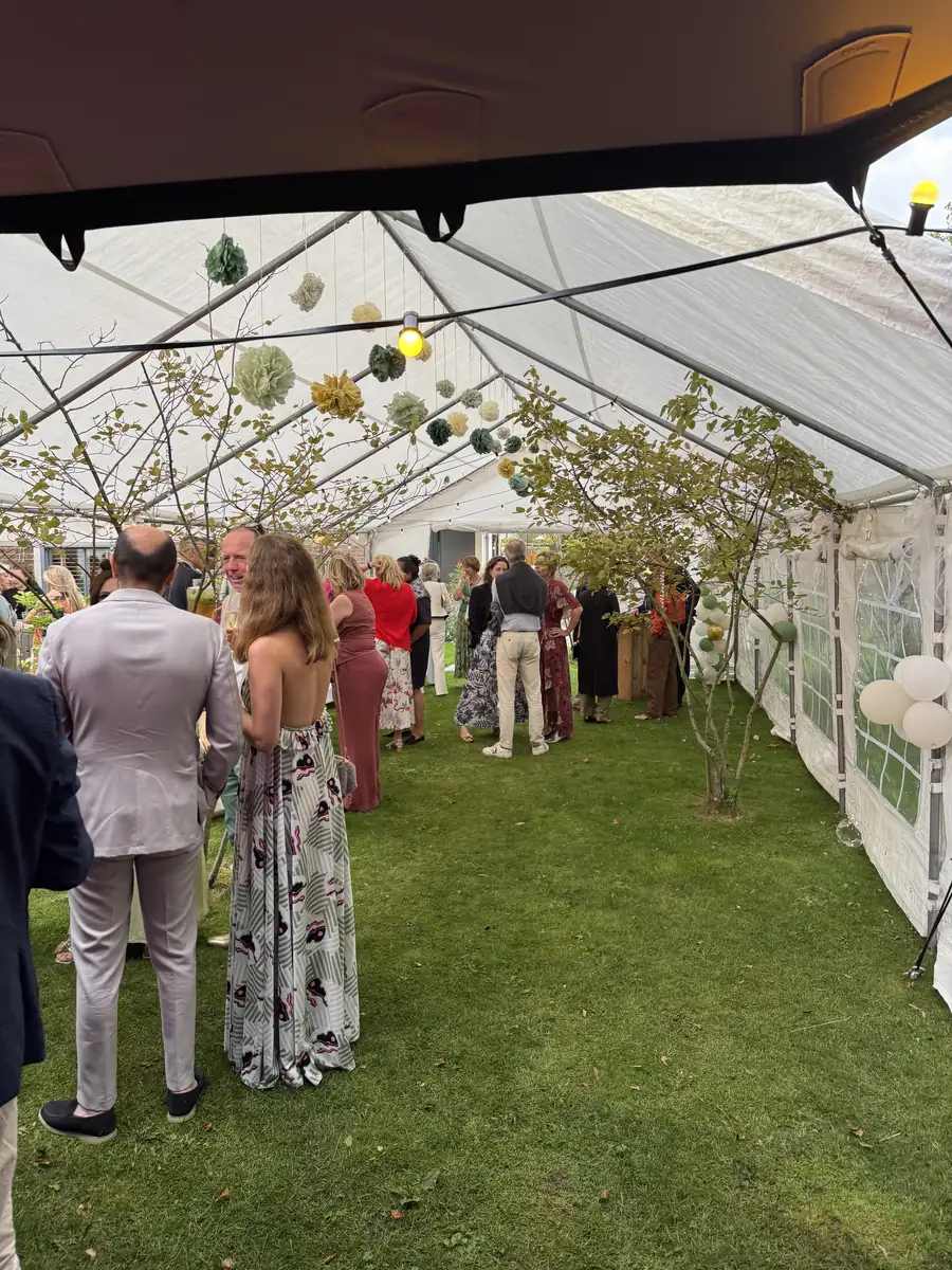 Gasten in feestkleding bij de borrel in een witte partytent op het gras, versierd met papieren pompons en lampjes, aan het begin van een bruiloftsfeest